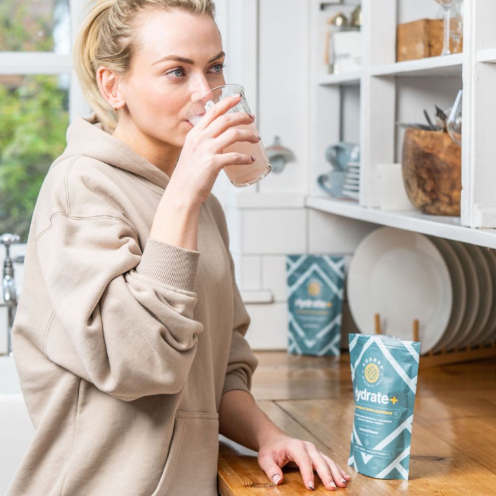 Junge, blonde Frau trinkt ein Glas Hydrate+ in ihrer Küche.