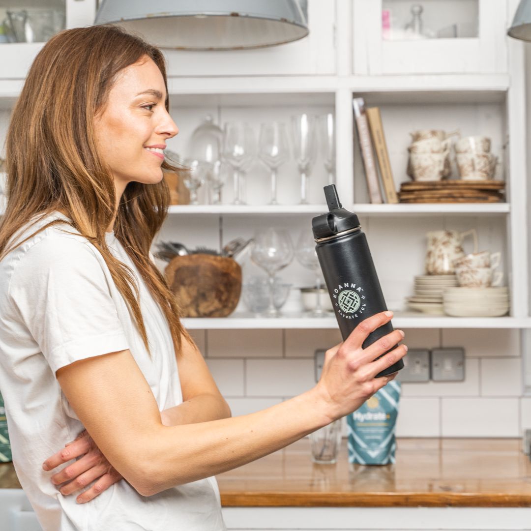 Junge Frau in ihrer Küche mit schwarzer Elevate Flasche in der Hand und Hydrate+ im Hintergrund.